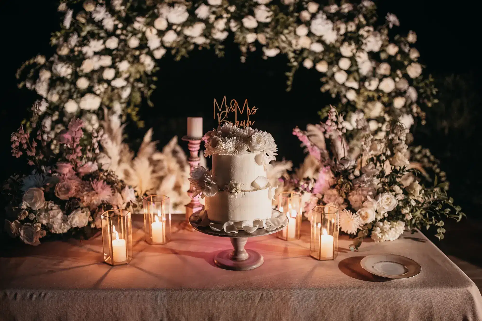 A candlelit cake table under an arch of white flowers