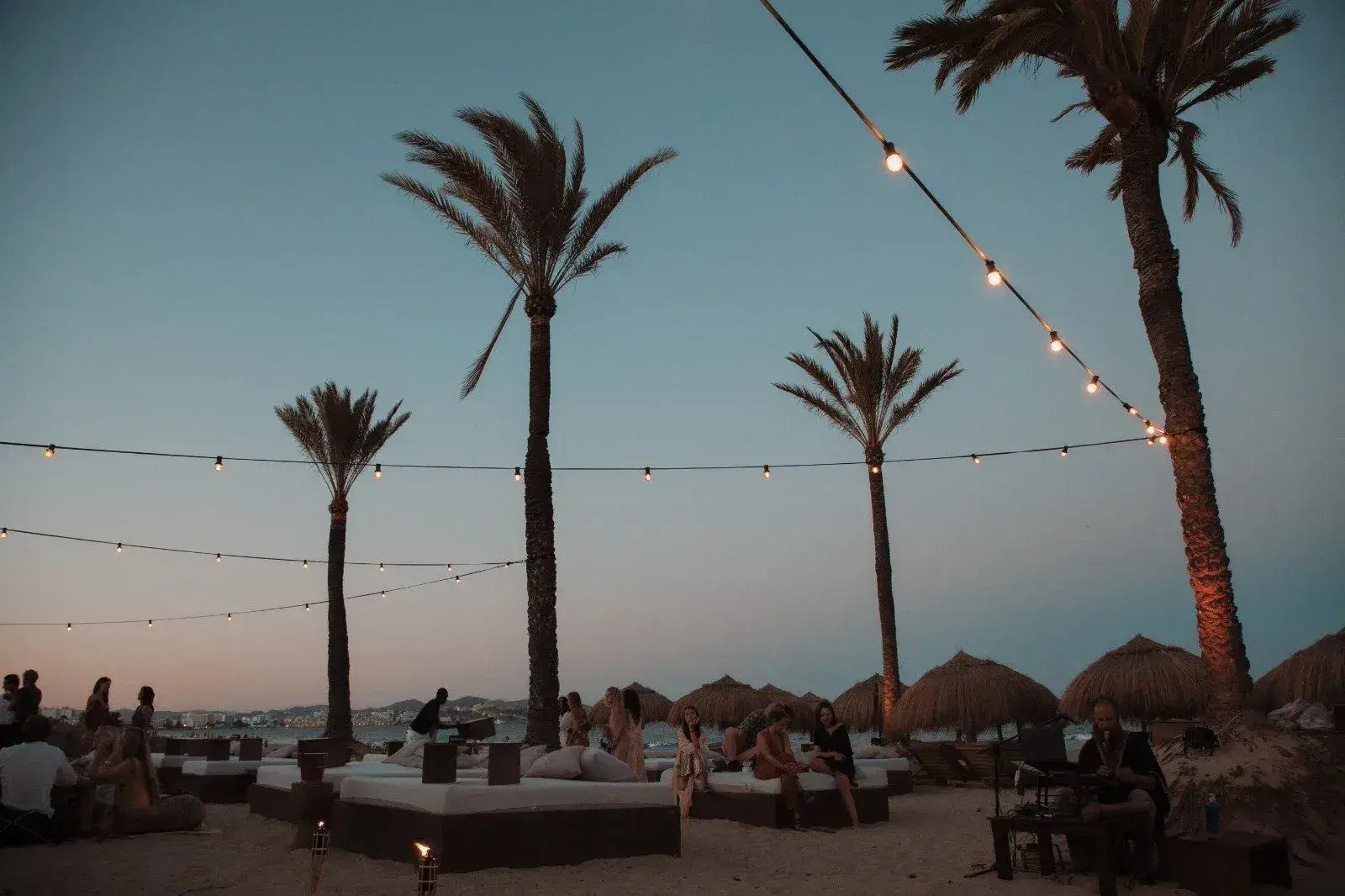 Beachouse Ibiza: Beach area at dusk under palms and festoon lights with guests at low tables