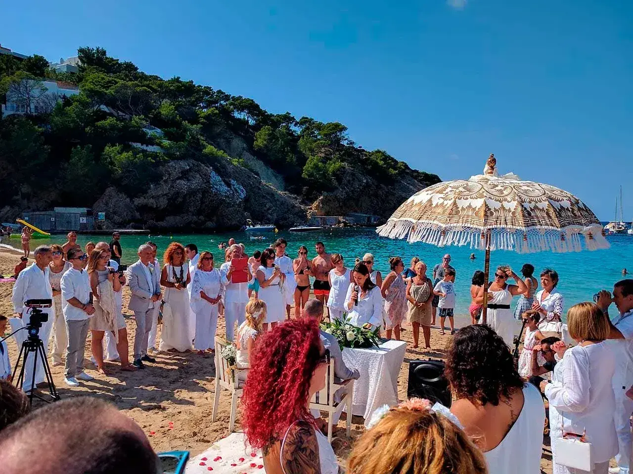 Cana Sofía: Wedding guests in white gathered around a beach ceremony under a fringed parasol on Cala Vadella
