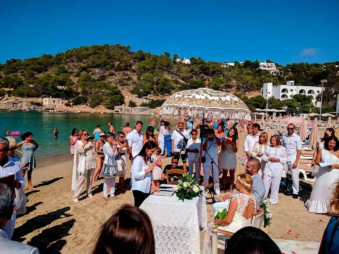 Cana Sofía: Beach ceremony with the couple at a table, guests standing around, and the bay behind