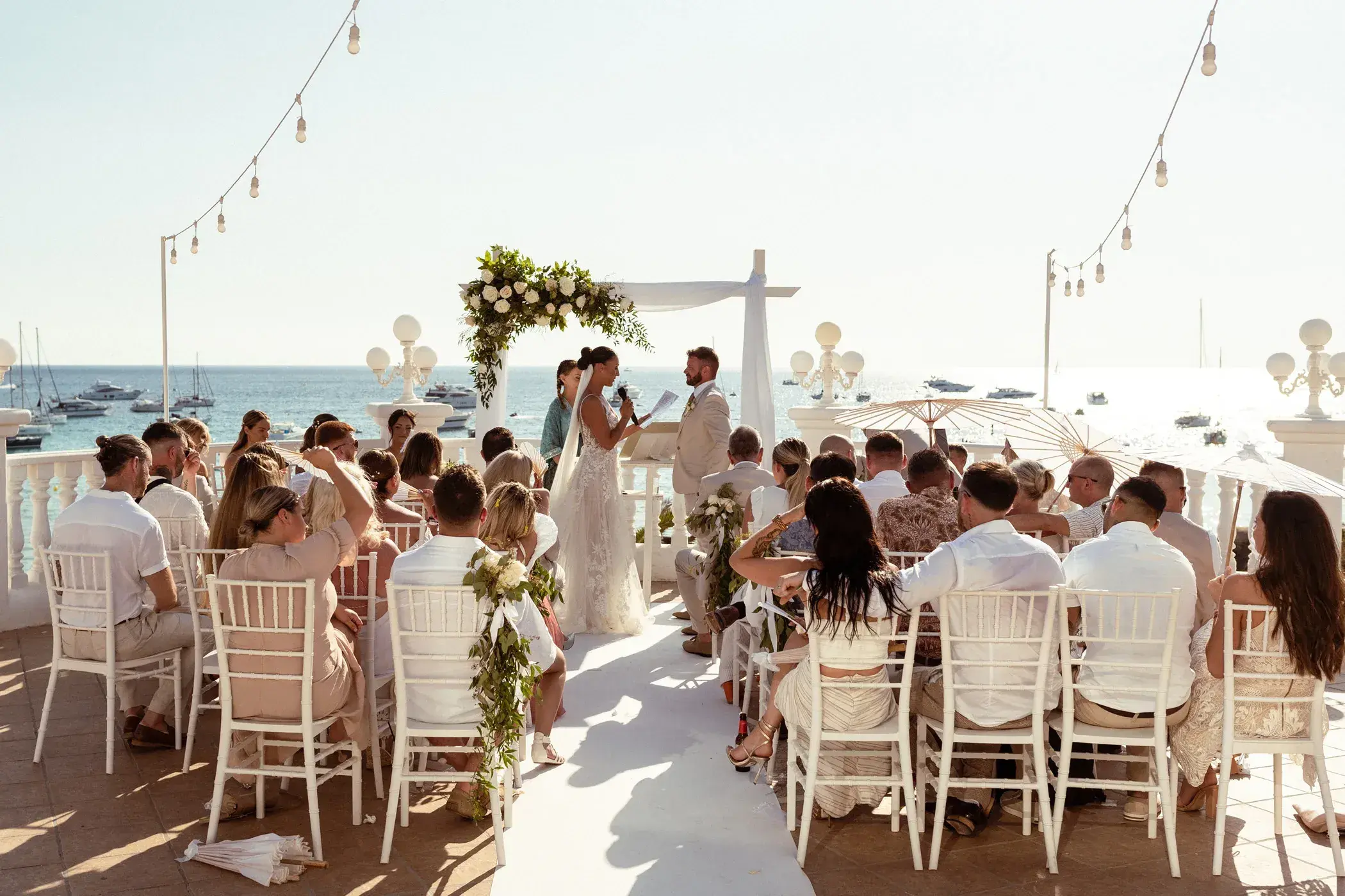 Cas Milà: Guests seated on white chairs for a ceremony on the terrace with festoon lights and the bay behind