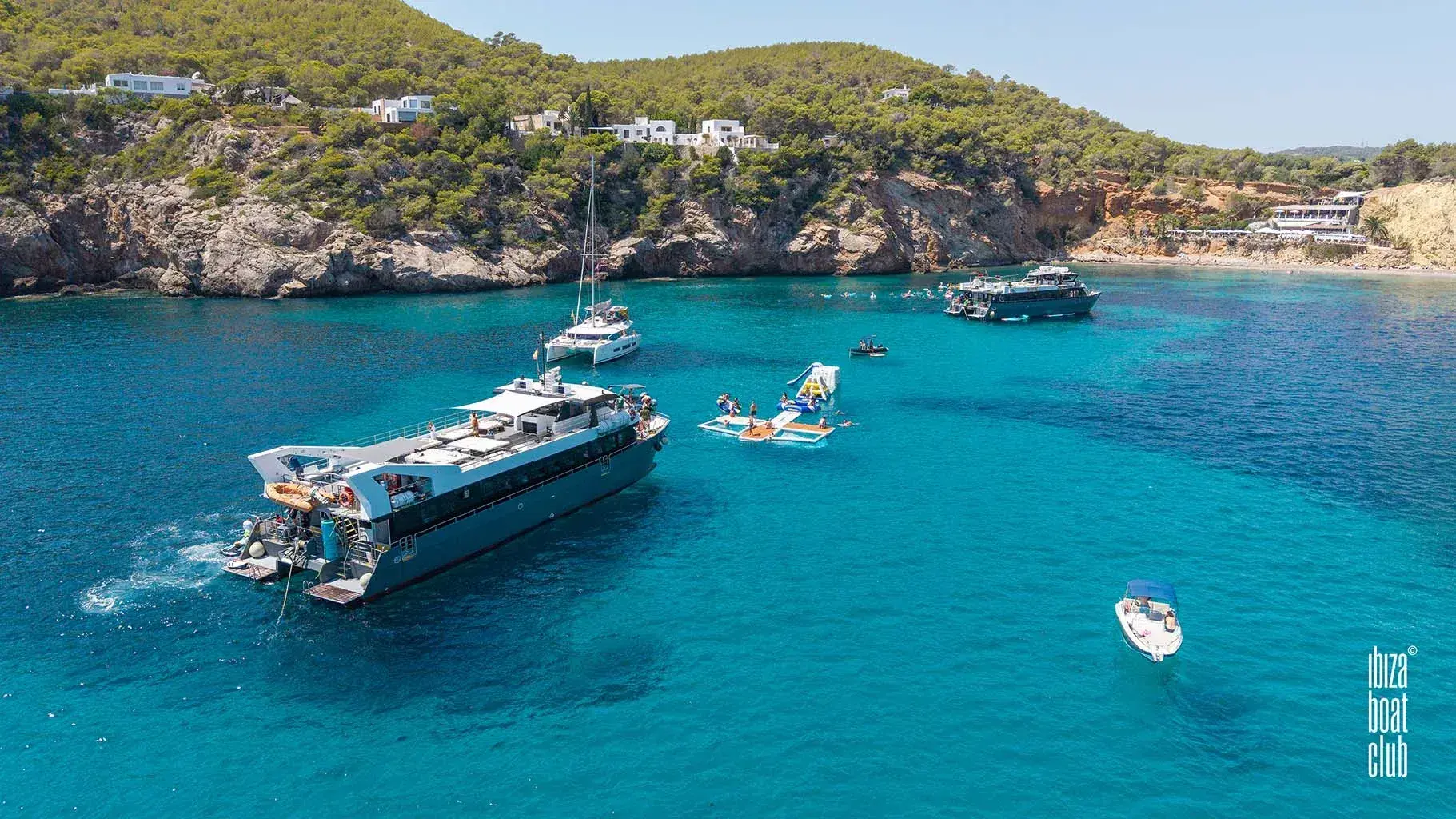 Ibiza Boat Club: The club boat anchored in a cove below pine-covered cliffs