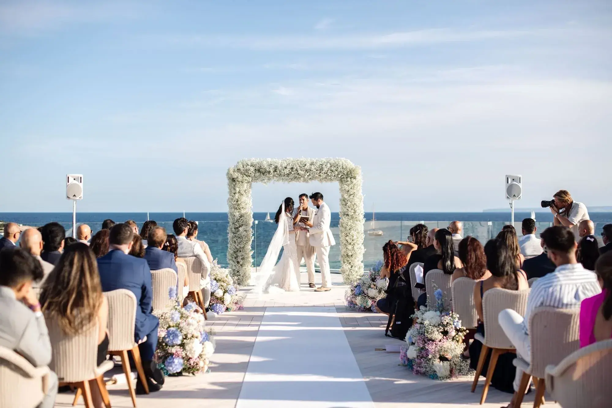 Nobu Hotel Ibiza Bay: Ceremony by the sea with guests seated either side of a white aisle and a floral arch